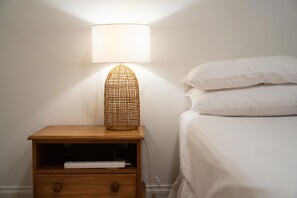 A wooden nightstand with a lamp and a drawer, next to a bed with white bedding. - Sunshine Daydream w Edmund Cottage (Auckland)