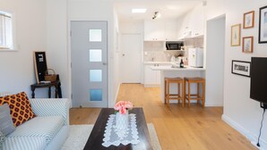 Ein modernes Wohnzimmer mit einer Couch, einem Couchtisch mit Blumen und einer Küche im Hintergrund. An der Wand ist ein Fernseher angebracht.
