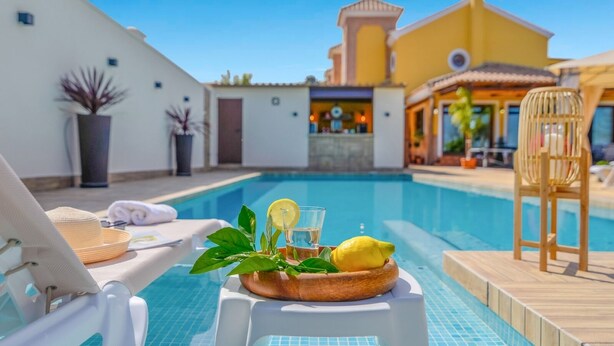 A poolside area with a lounge chair, a small table with a drink and lemon, and a view of a building with a yellow facade. - Residencial Las Torrecillas, Casa 15 Puente Genil by Ruralidays (Córdoba)