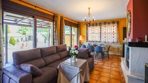 A cozy living room with a sofa, a dining area with chairs, a chandelier, and large windows with curtains. - Residencial Las Torrecillas, Casa 15 Puente Genil by Ruralidays (Córdoba)