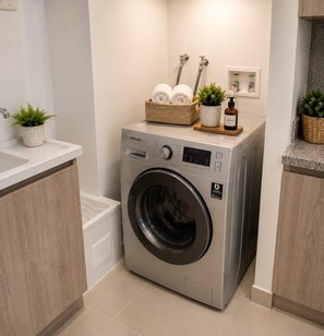 A modern laundry room with a front-loading washing machine, a sink, and a small shelf with towels and plants.