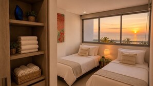 Een hotelkamer met twee bedden, een groot raam met uitzicht op een zonsondergang, een houten plank met handdoeken en een tafeltje met een lamp.