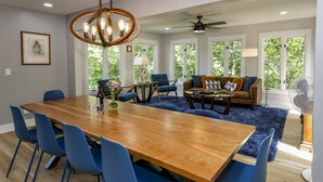 Une salle à manger spacieuse avec une table en bois et des chaises bleues, un salon avec un canapé et des fauteuils, et de grandes fenêtres donnant sur un paysage verdoyant.