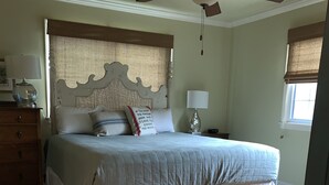 A bedroom with a bed, wooden floor, ceiling fan, dresser, nightstand, lamp, and window with blinds. - Walk  to beach & 7 restaurants.  Our 1950’s 3 bed & 2 bath cottage is fantastic! (St. Simons)