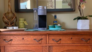 A wooden cabinet with drawers, a lamp, a Keurig coffee maker, a jar of biscotti, a bottle of Torani syrup, and a potted orchid. - Walk  to beach & 7 restaurants.  Our 1950’s 3 bed & 2 bath cottage is fantastic! (St. Simons)