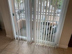 A room with a balcony featuring white blinds, a tiled floor, and a view of a parked car and greenery outside.