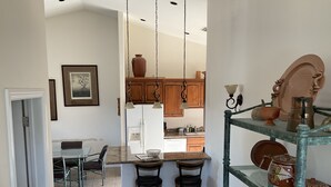 Een keuken met houten kastjes, een witte koelkast en een hangende lamp. Er is een eethoek met een tafel en stoelen, en een bar met krukken.