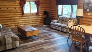 L’intérieur d’une cabane en rondins, avec un canapé, une table basse en bois, une table à manger avec des chaises, une cheminée et un escalier.