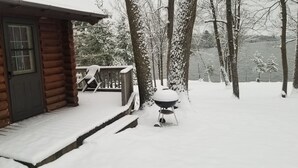 Une cabane en rondins avec une terrasse en bois et un barbecue recouvert de neige.