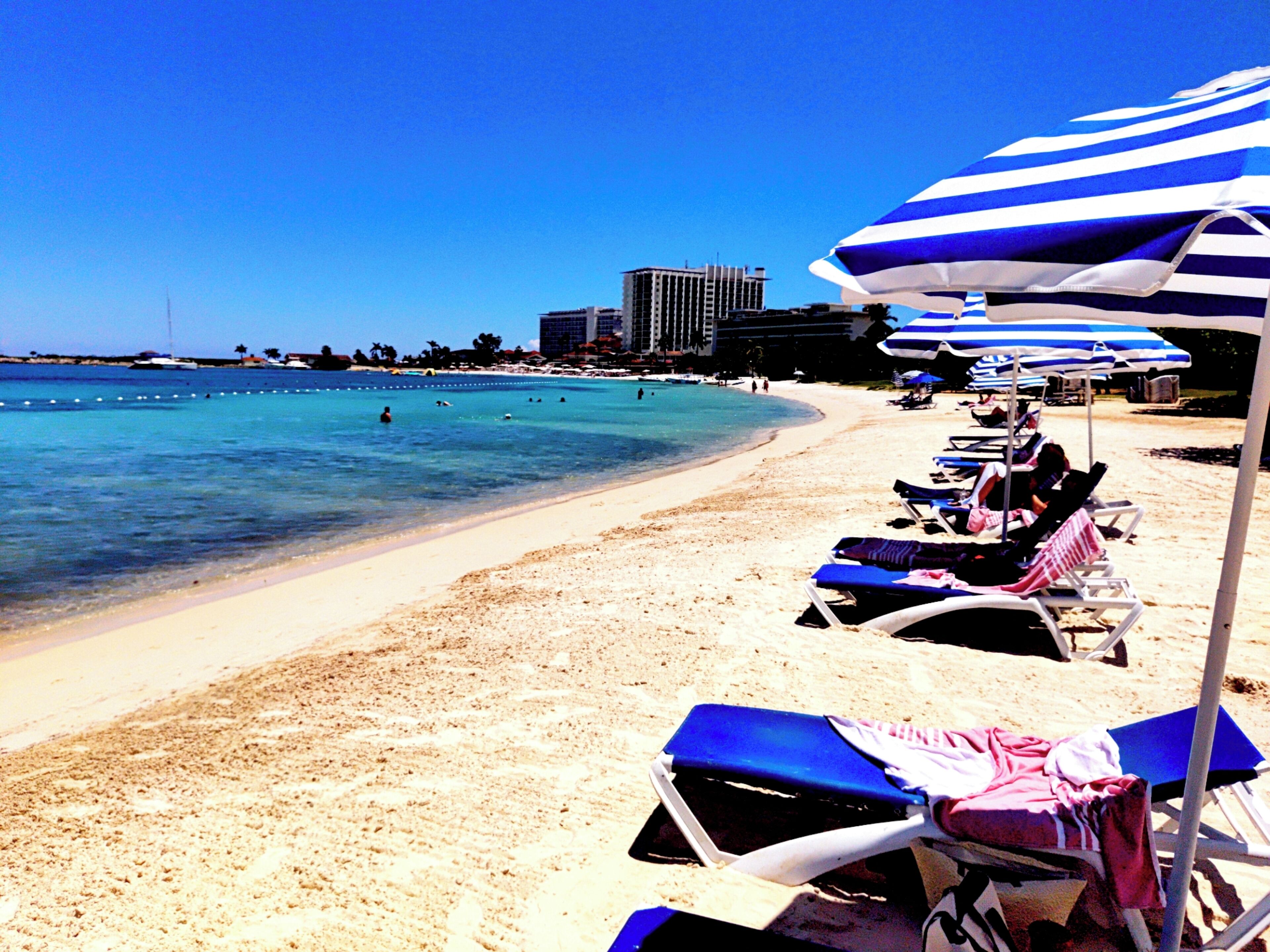 A beach with rows of sun loungers and umbrellas, a clear blue sea, and buildings in the background.