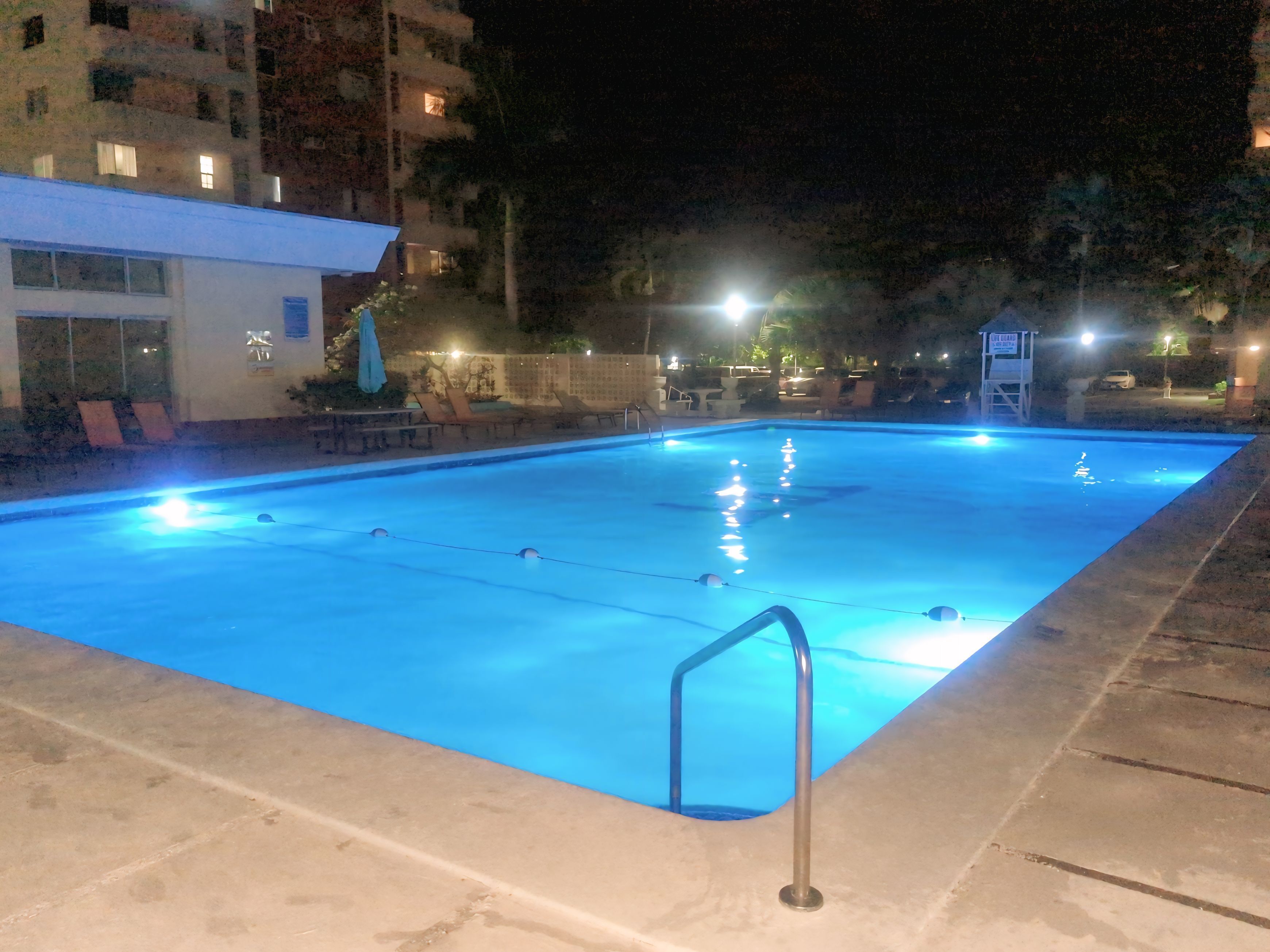 A nighttime swimming pool with clear water, surrounded by a low wall and a few lounge chairs.