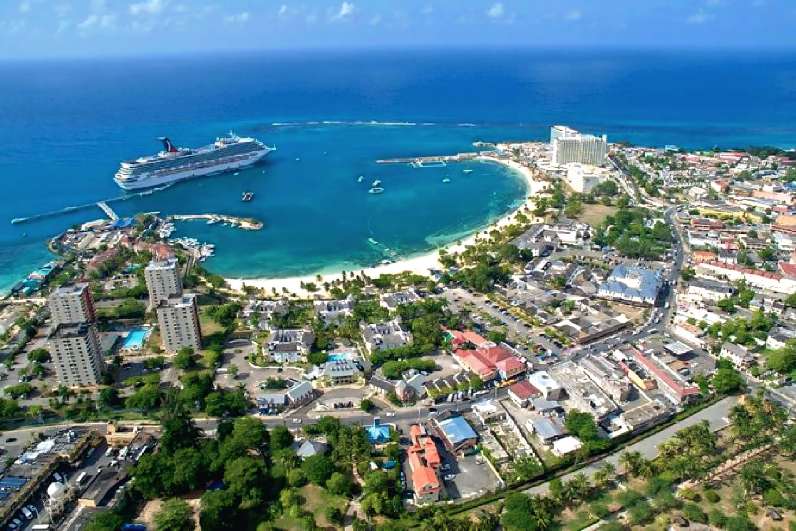Un paisaje urbano costero con un crucero amarrado cerca de una playa, rodeado de edificios residenciales y comerciales.