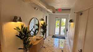 A hallway with marble flooring, a wooden bench, potted plants, wall-mounted lighting, and a door leading outside.