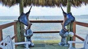 A thatched-roof structure with two metal sculptures of fish on a wooden deck overlooking the sea.