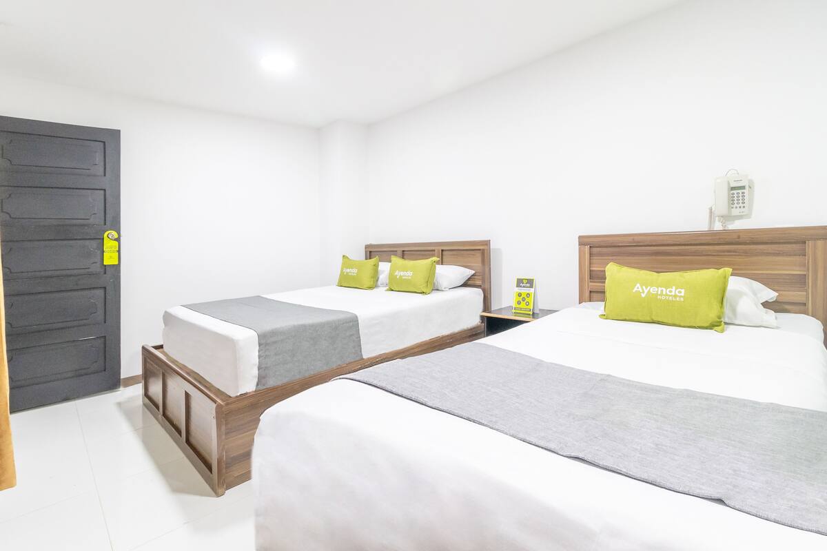 A hotel room with two beds, each with a wooden headboard, grey and white bedding, and green pillows with the brand 'Ayenda'.