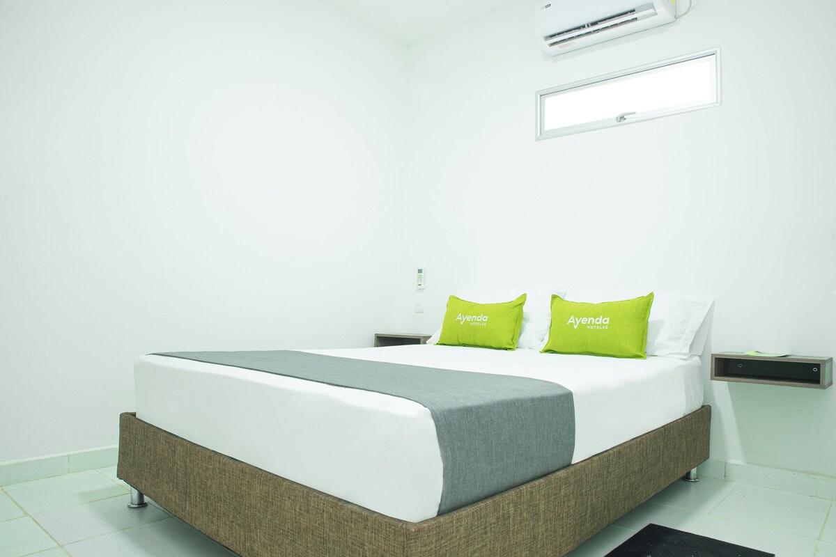 Una habitación de hotel moderna con una cama grande, ropa de cama gris y blanca, y almohadas verdes con la marca 'Ayenda Hotels'.