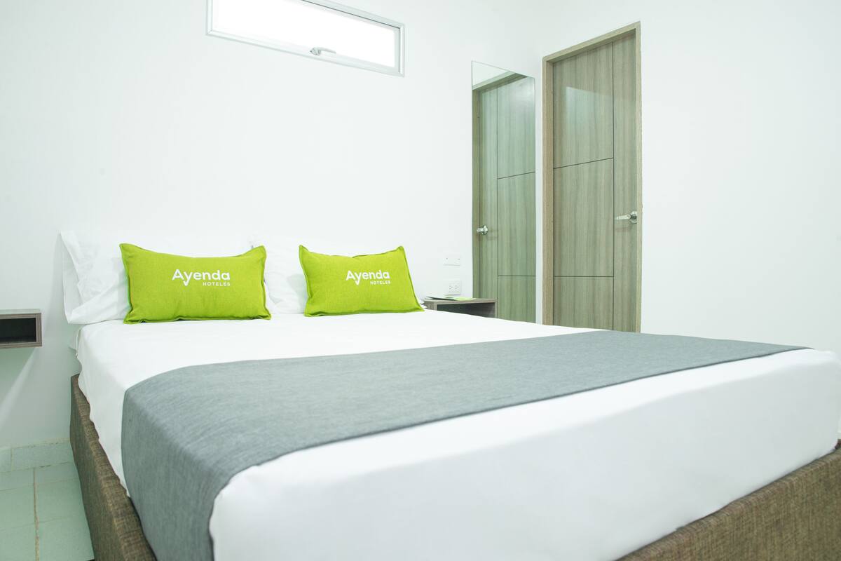 Una habitación de hotel con una cama, dos almohadas verdes, una manta gris y un armario de madera.