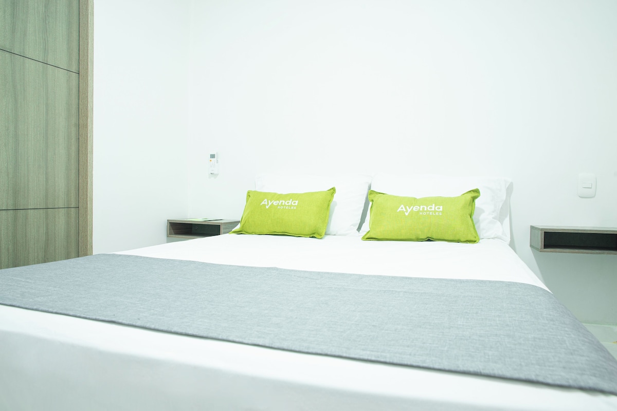 Una habitación de hotel con una cama, dos almohadas con el nombre del hotel, un edredón gris y un armario de madera.