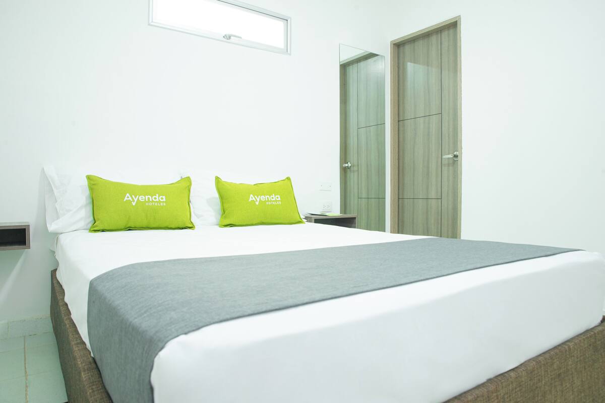 Una habitación de hotel con una cama, dos almohadas verdes, una manta gris y un armario de madera.