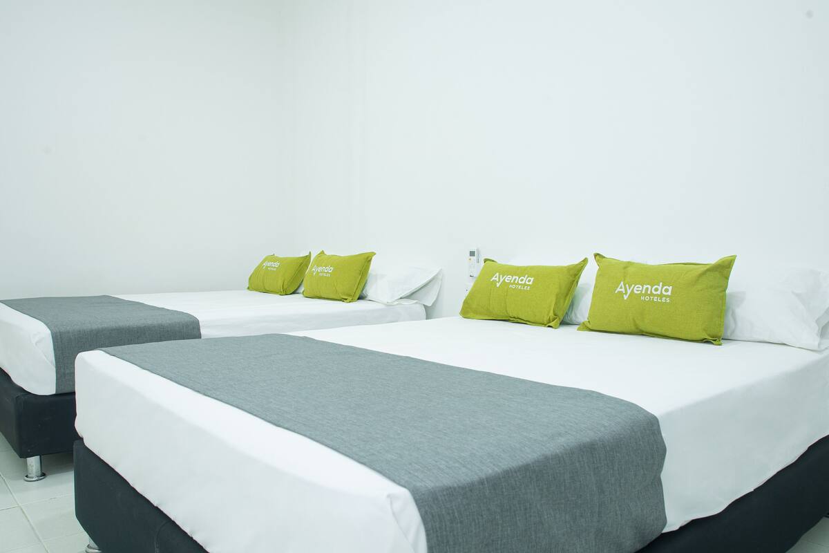 Habitación de hotel con dos camas individuales, cada una con una colcha gris y una almohada verde con la etiqueta "Ayenda Hotels".