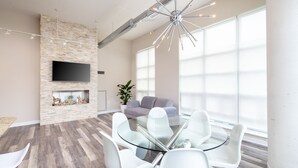 Una sala de estar moderna con una pared de piedra, un televisor de pantalla plana, una chimenea, un ventanal grande y una mesa de comedor de vidrio con sillas.