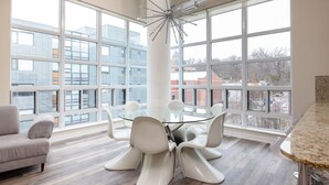 Un comedor moderno con una mesa de vidrio y sillas blancas, ventanas amplias con vistas a la ciudad y una lámpara de techo original.