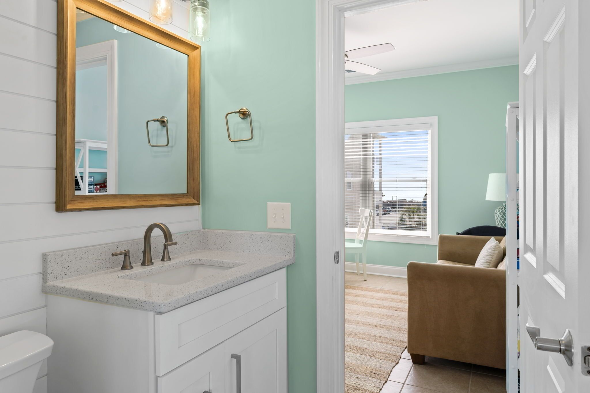 A bathroom with a white sink, a large mirror, and a door leading to a living area with a beige sofa and a window with blinds.