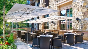 An outdoor patio area with tables and chairs, stone walls, and large umbrellas providing shade.