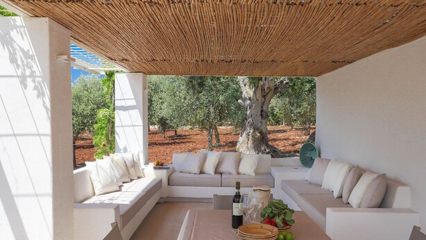 Zona relax all'aperto coperta, con un divano, un tavolo con una bottiglia, bicchieri e frutta.