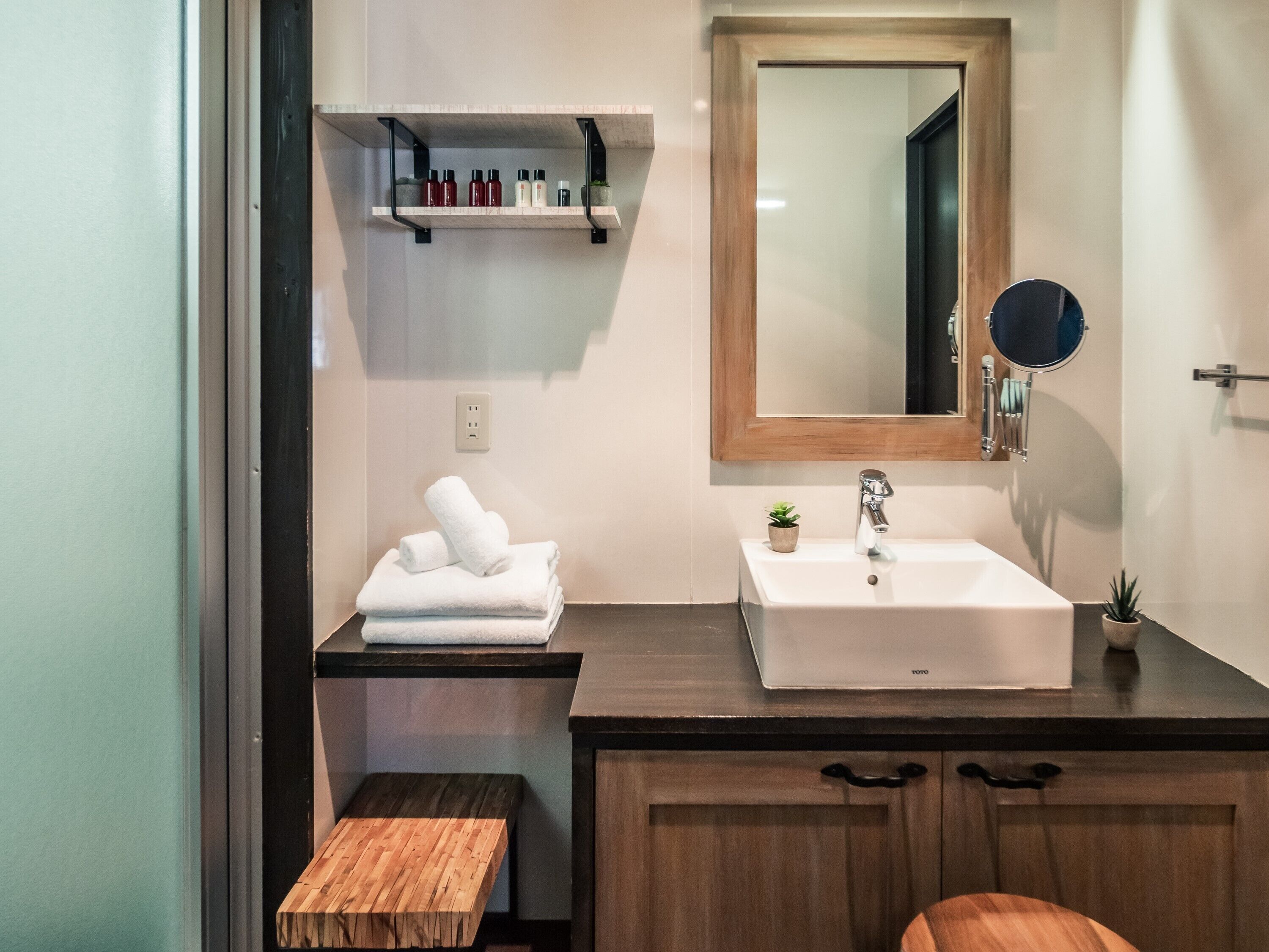 A bathroom with a white sink, wooden vanity, a mirror, a shelf with toiletries, and a small bench.