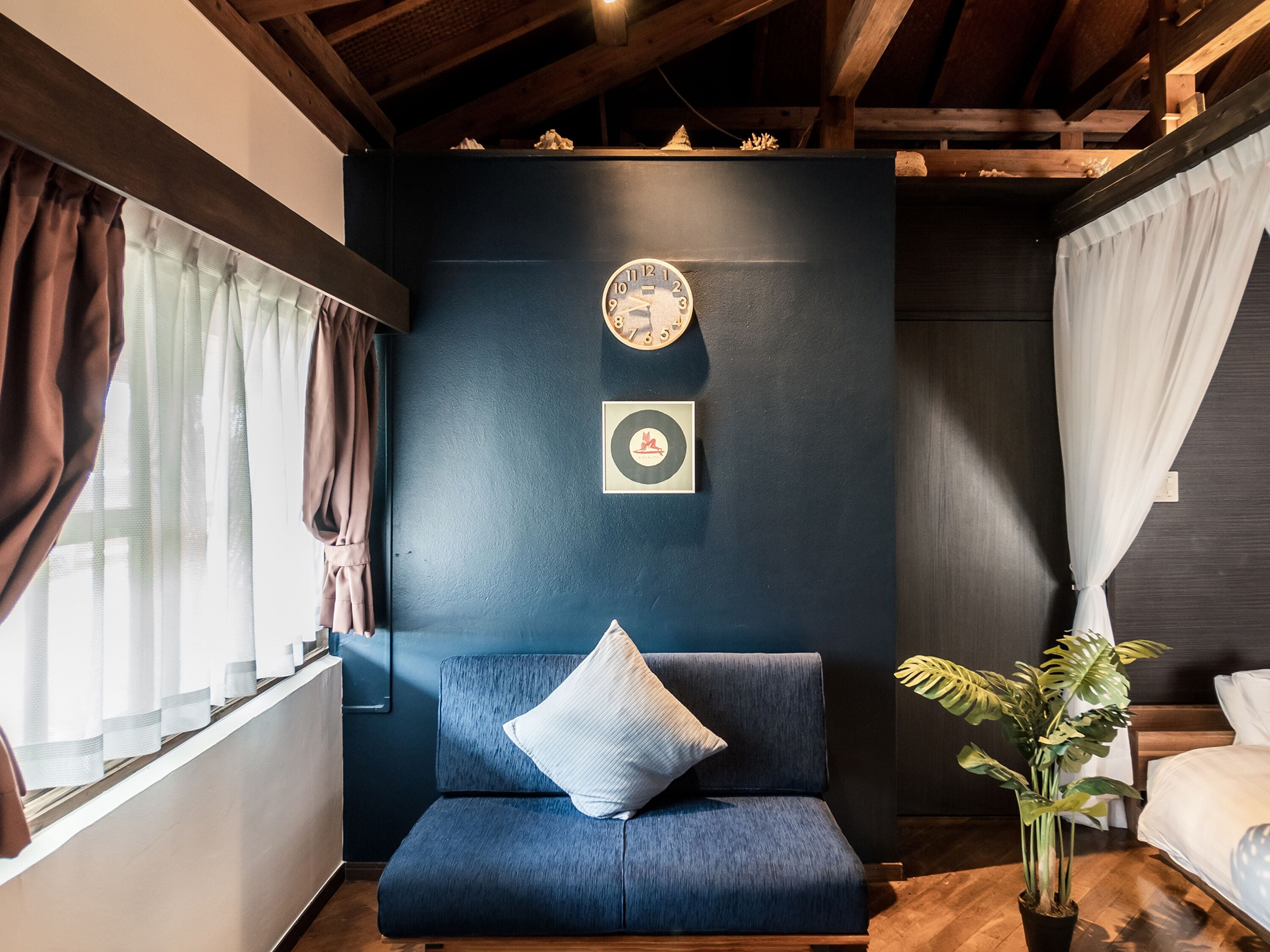 A room with a dark blue accent wall, a clock, a framed picture, a blue sofa with a pillow, and a potted plant.