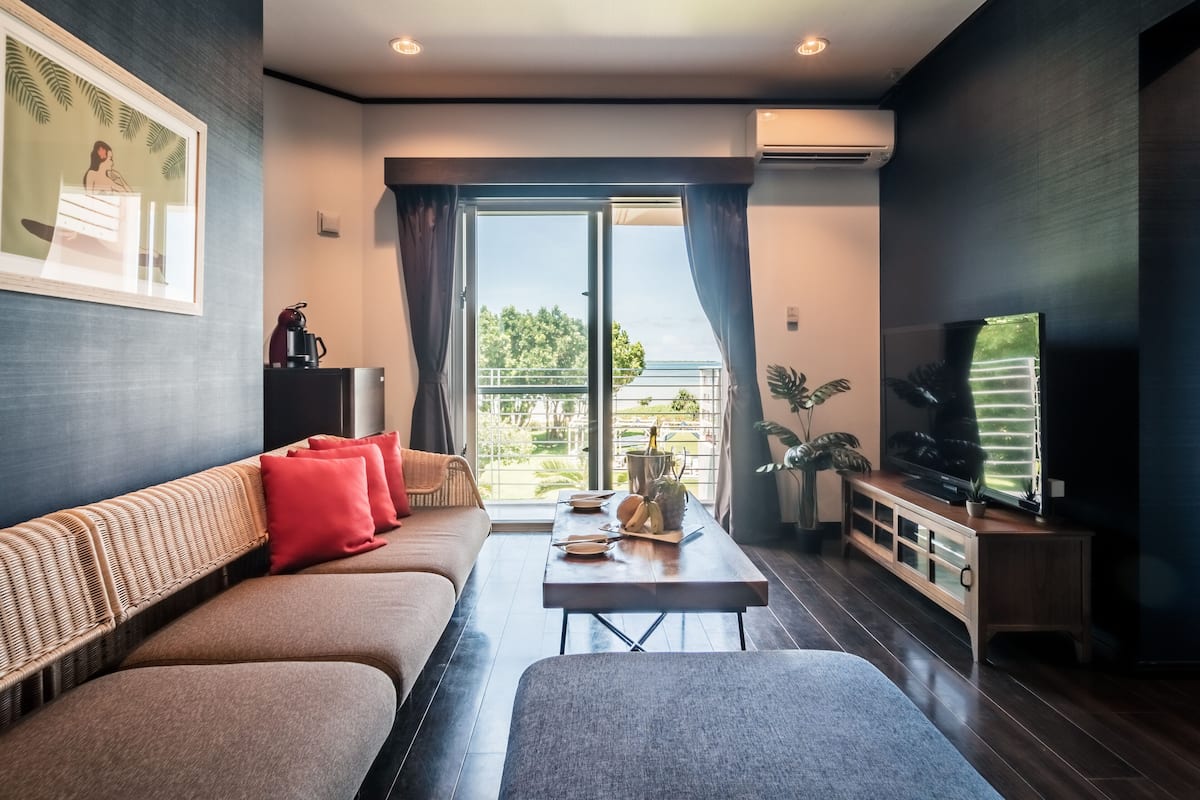A modern living room with a sofa, a coffee table, a TV, and a balcony with a view of the outdoors.