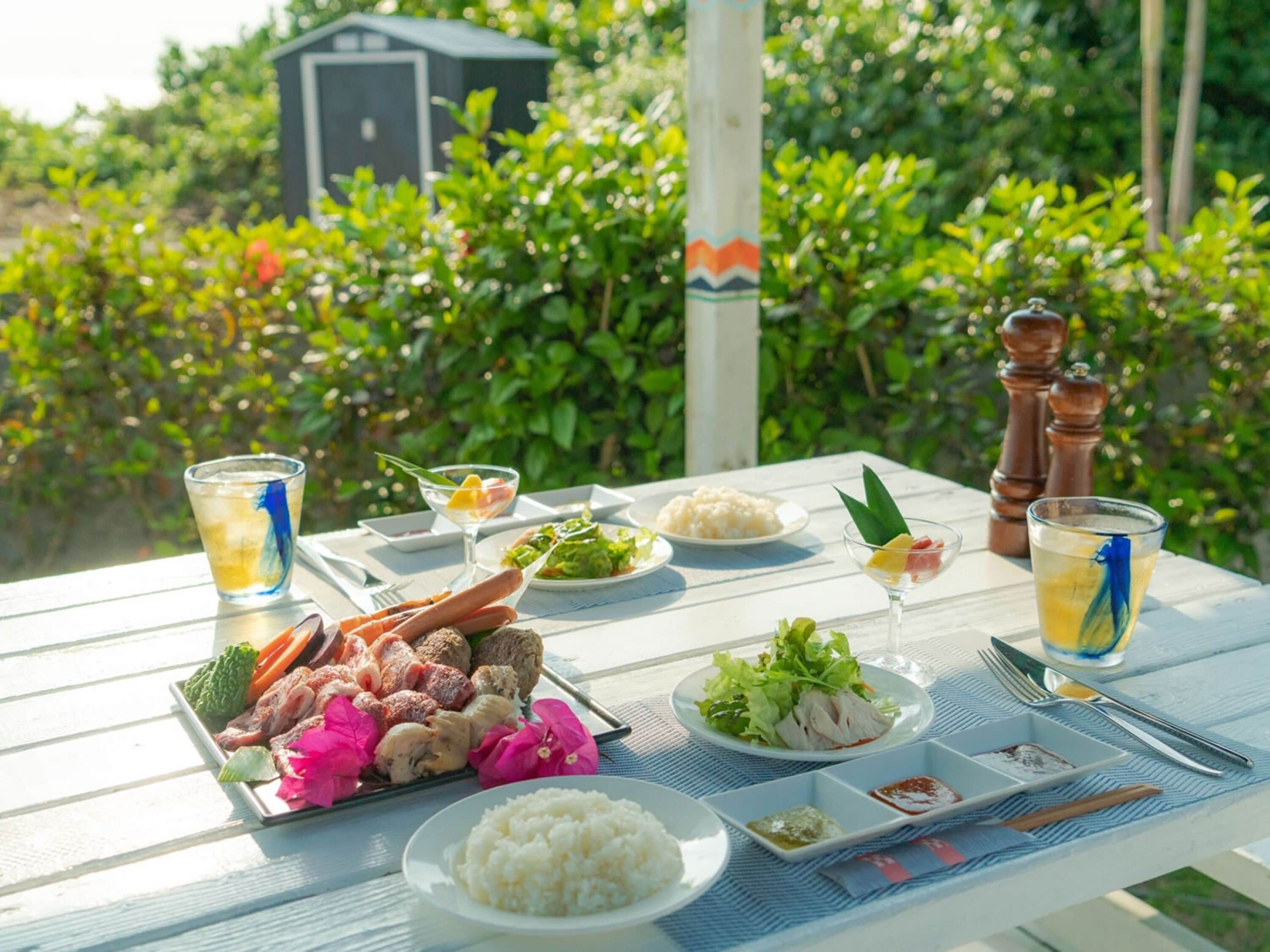 A table set with various dishes, drinks, and condiments, surrounded by lush greenery.