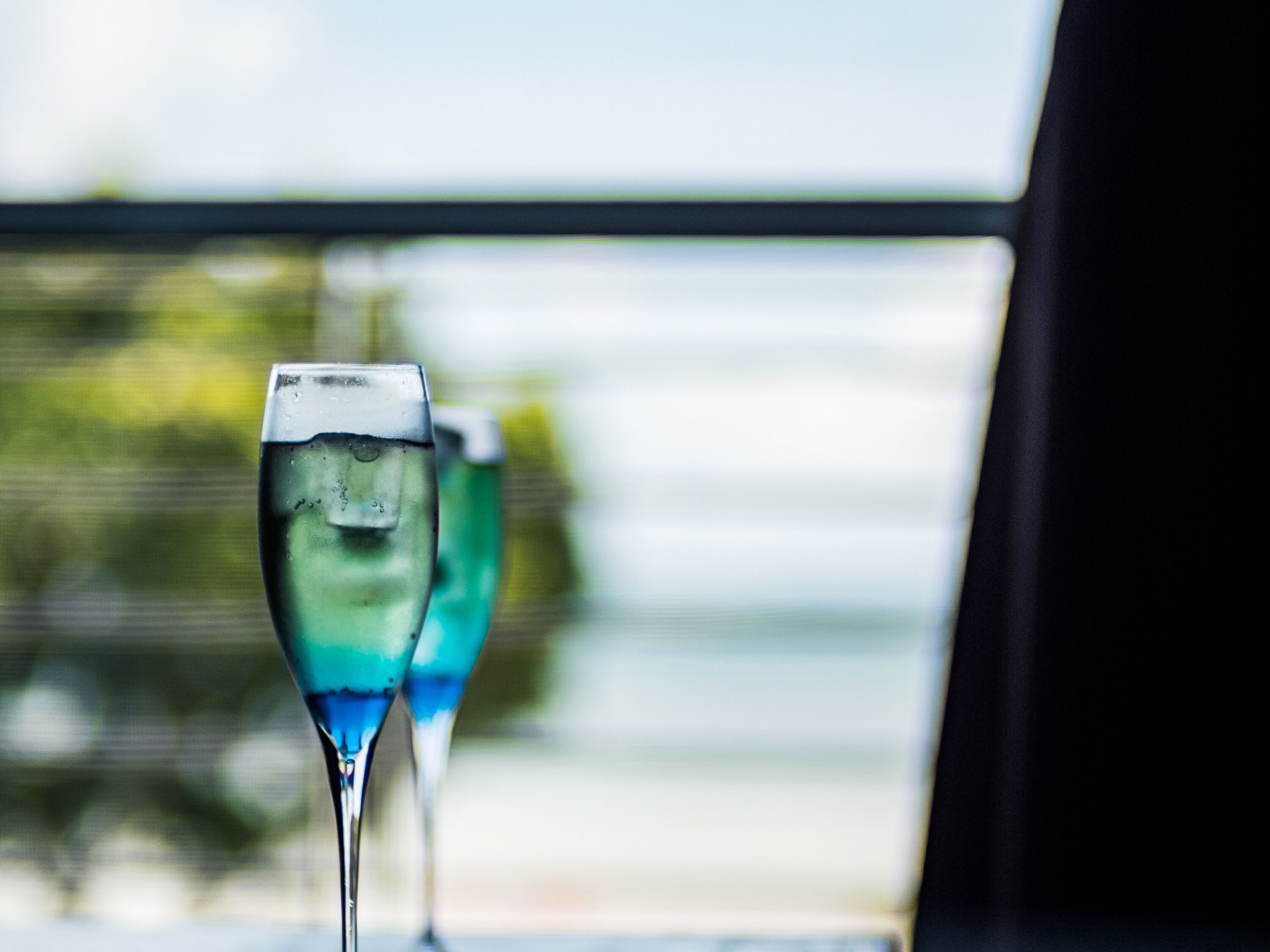 Two glasses of blue liquid on a table with a blurred background of greenery and a window.