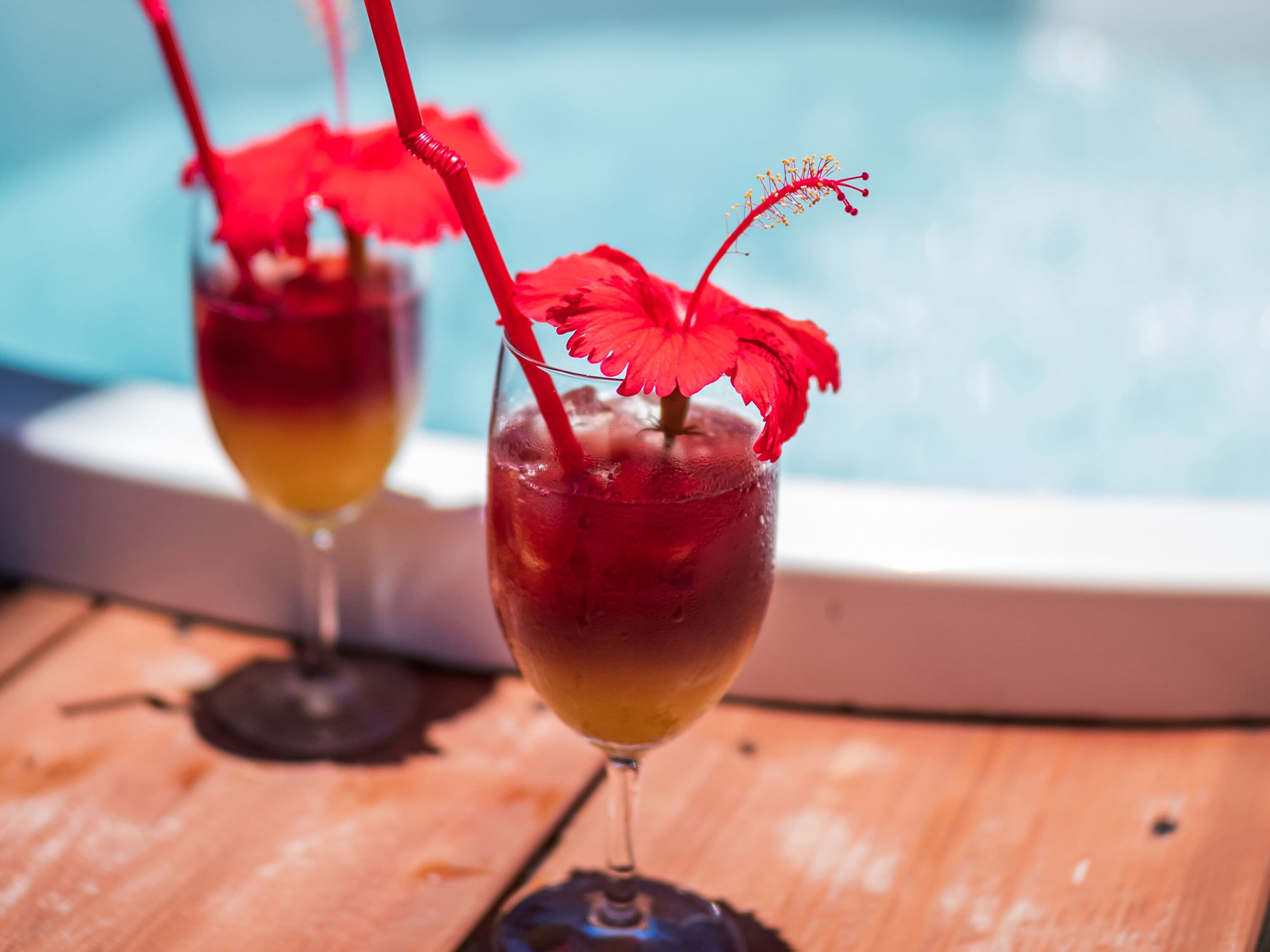 Two cocktails with red straws and a red flower garnish, placed on a wooden table near a pool.