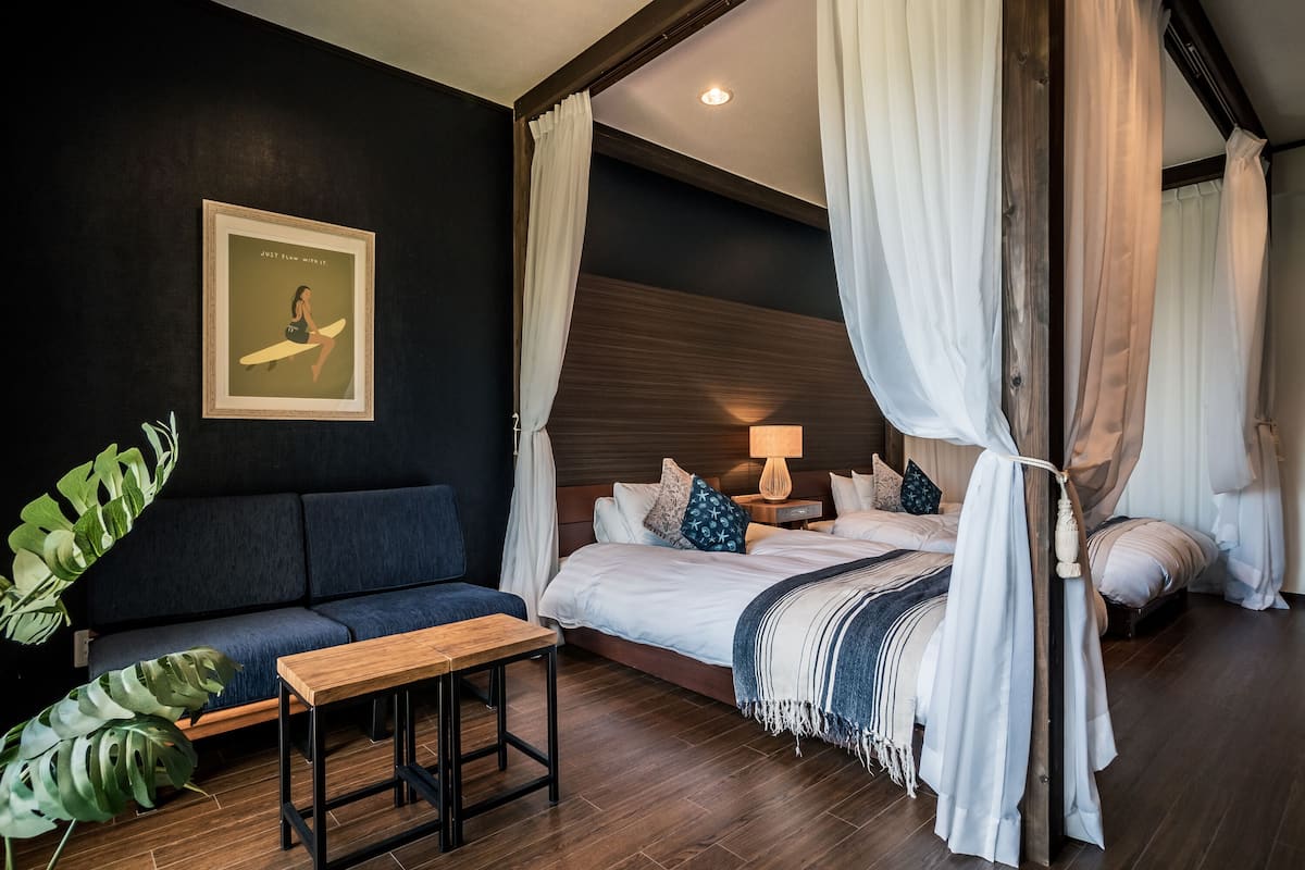 A bedroom with a bed, a bench, a nightstand, a lamp, and a large plant.