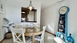 A dining area with a round table, white chairs, a hanging light fixture, a wall clock, and a bookshelf.