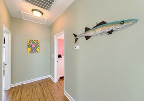 A hallway with a wooden floor, a ceiling vent, a wall-mounted fish decoration, and a colorful painting. - Billy-Lisa - Second Row in Garden City Beach, SC (Garden City)