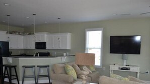 A modern kitchen with white cabinets, a central island with stools, a flat-screen TV, and a ceiling fan. - Billy-Lisa - Second Row in Garden City Beach, SC (Garden City)
