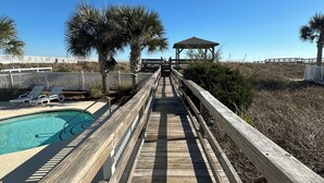 Une promenade en bois mène à une zone de plage équipée d’une piscine et de chaises longues.