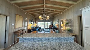 Une pièce spacieuse avec un plafond haut, des poutres en bois, un grand lustre, un coin repas doté d’un comptoir en granit et un coin salon confortable avec un canapé et des fauteuils.