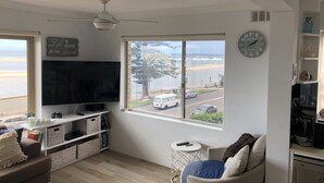 Una sala de estar con televisor, un ventanal con vista a la playa, un ventilador de techo y un reloj en la pared.