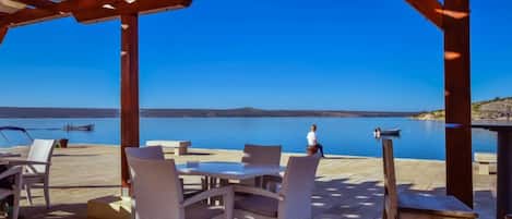 Un área de estar al aire libre con sillas y mesas de mimbre, con vista a un cuerpo de agua tranquilo y una costa lejana.