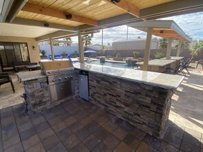 Cocina exterior con encimera de piedra, parrilla empotrada y vistas a un patio trasero con piscina y mobiliario de jardín.