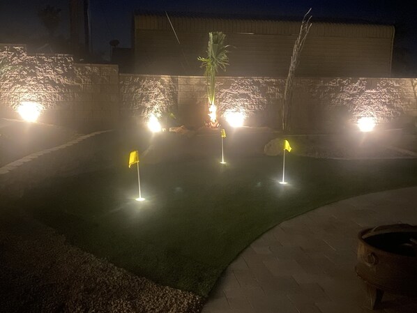 Vista nocturna de un área ajardinada con iluminación artificial, una palmera central y tres banderas iluminadas.