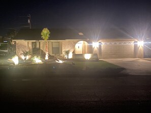 Una casa residencial con garaje, iluminada por luces exteriores y un césped bien cuidado con plantas pequeñas.