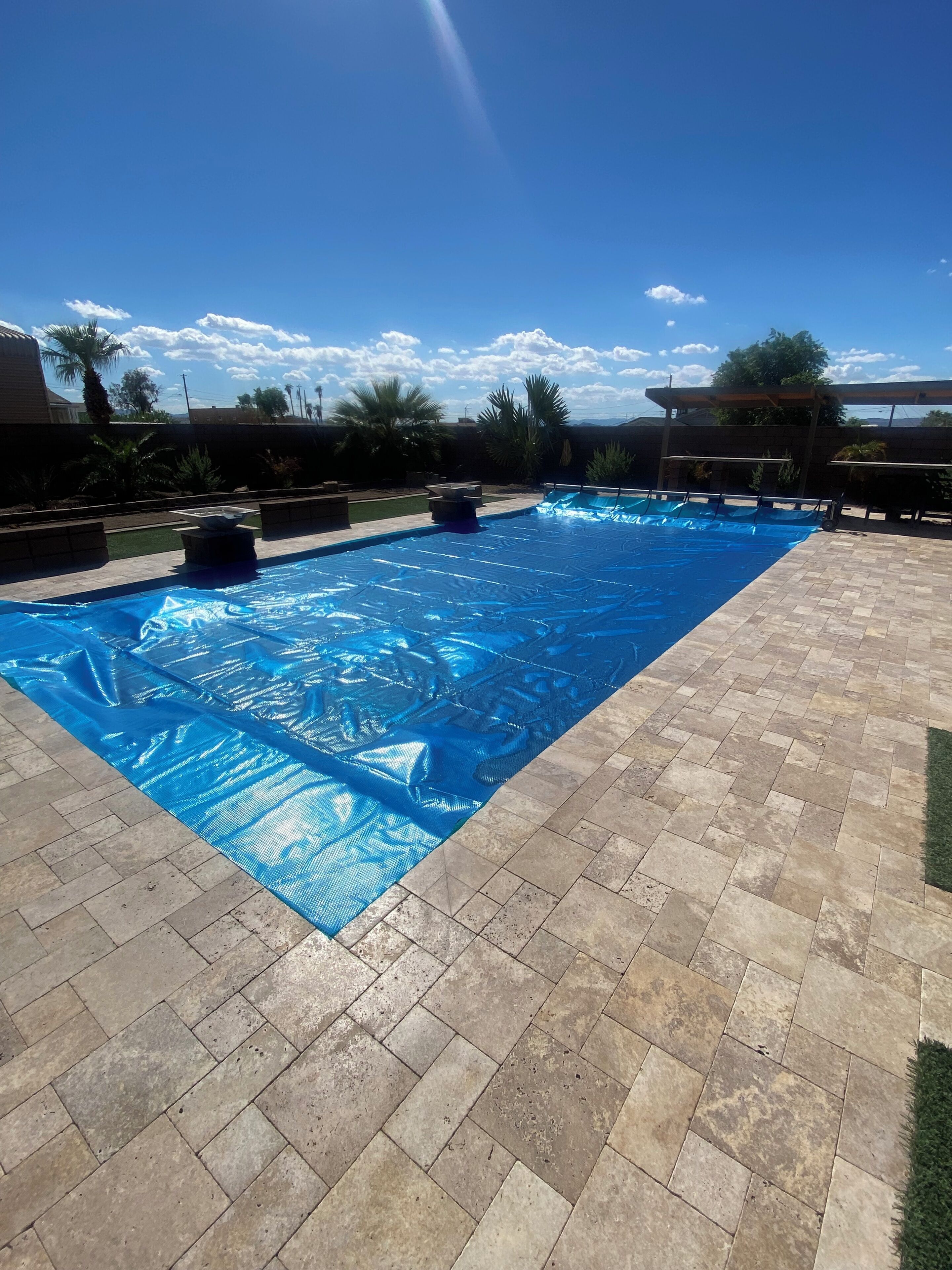 Piscina rectangular con cubierta azul, rodeada por un patio de ladrillo y palmeras.