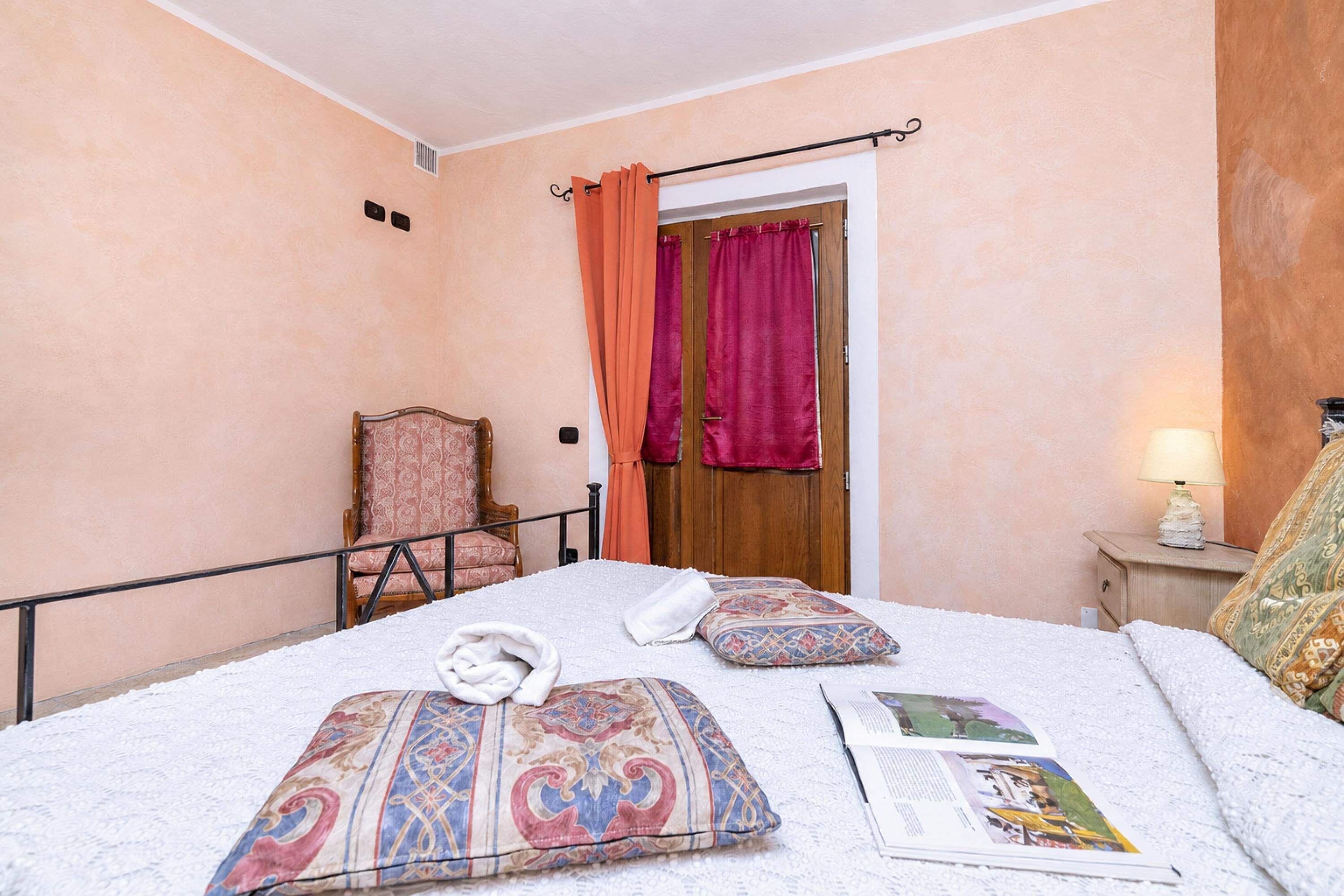 Un dormitorio con cama, almohadas, una silla, una puerta y una mesita de noche con lámpara.