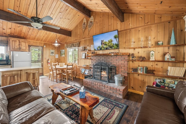Interior de una cabaña de madera con chimenea, mobiliario de cuero, televisor y zona de cocina.