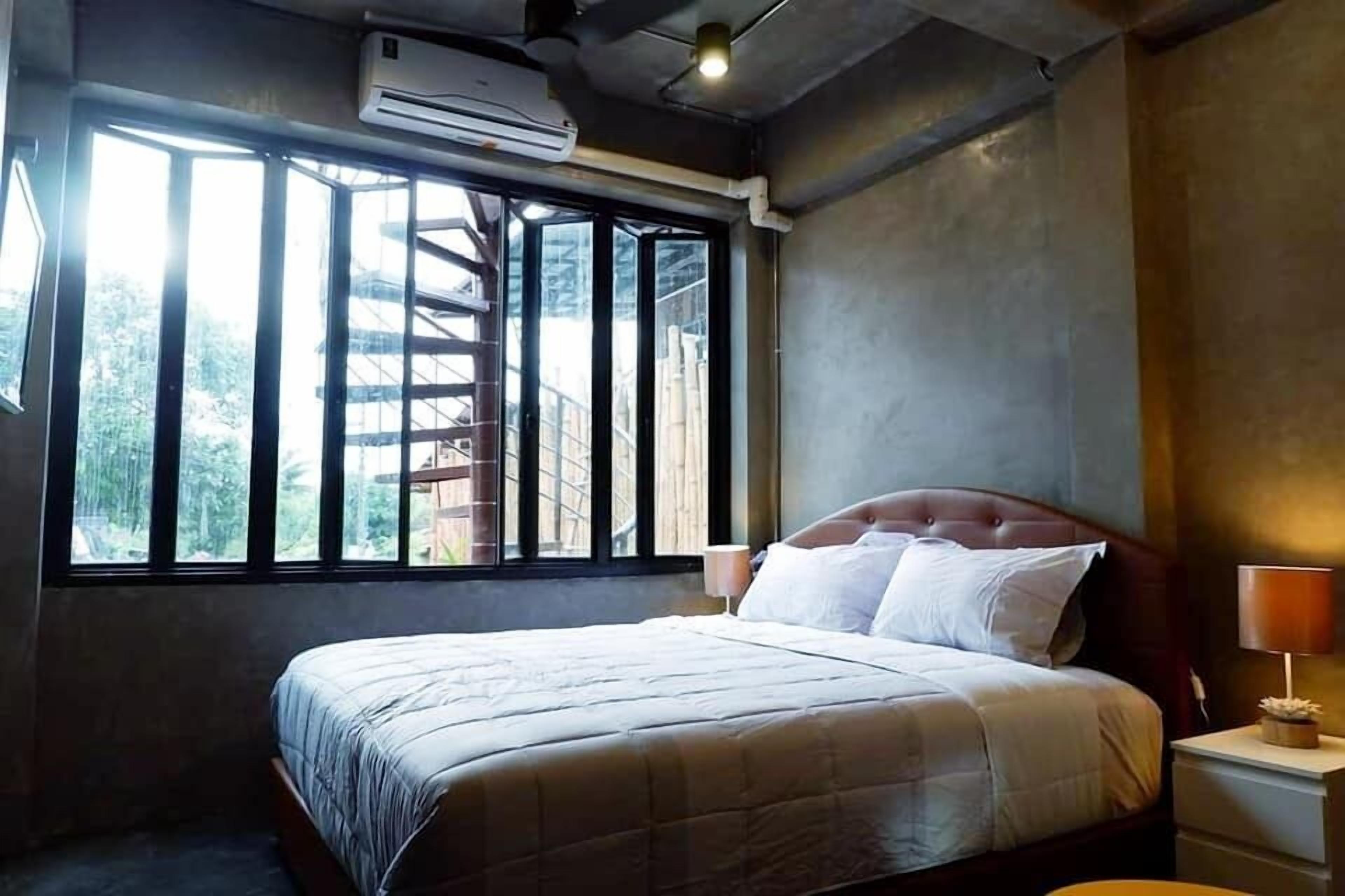 A bedroom with a bed, a nightstand, an air conditioning unit, and a window with a view of a staircase and greenery outside.